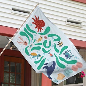 May include: A white flag with a colorful ocean-themed design. The flag features illustrations of sea creatures like an octopus, whale, seahorse, and crab, along with seaweed and bubbles. The flag is displayed outside a house.