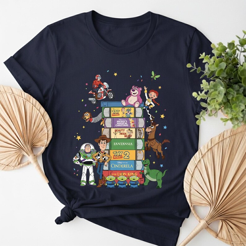 Może przedstawiać: Granatowa koszulka z kolorową grafiką postaci Disneya i tytułami film&oacute;w na stosach książek. Projekt zawiera postacie z "Toy Story", "Pięknej i Bestii" oraz "Kopciuszka".