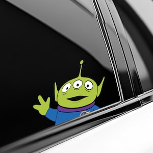 May include: A green alien sticker with three eyes, a blue shirt, and a purple collar. The alien is waving with a yellow hand. The sticker is applied to a car window.