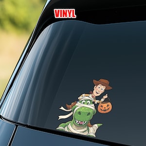 Puede incluir: Una colorida calcoman&iacute;a de vinilo con Woody de Toy Story montado en un dinosaurio momificado, sosteniendo una linterna de calabaza. La palabra "VINYL" est&aacute; en texto rojo y blanco en la parte superior. La calcoman&iacute;a est&aacute; dise&ntilde;ada para una ventana de coche.