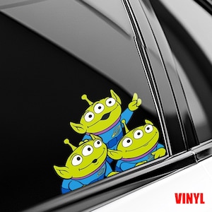 May include: A vinyl decal featuring three green cartoon aliens with large eyes, wearing blue uniforms. One alien is pointing upwards. The word "VINYL" is printed in red at the bottom right.