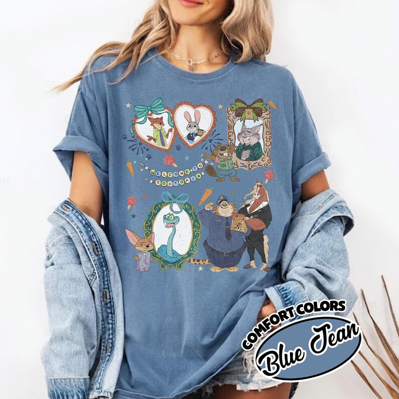 May include: A blue jean colored t-shirt featuring a graphic print of various animated animal characters from Zootopia. The design includes framed portraits, a banner with the text Welcome to Zootopia, and scattered carrot and popsicle icons.
