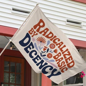 May include: A cream-coloured flag displaying the text "RADICALIZED BY BASIC DECENCY" in orange and navy blue, with floral designs. The flag is mounted outside a house with red trim.