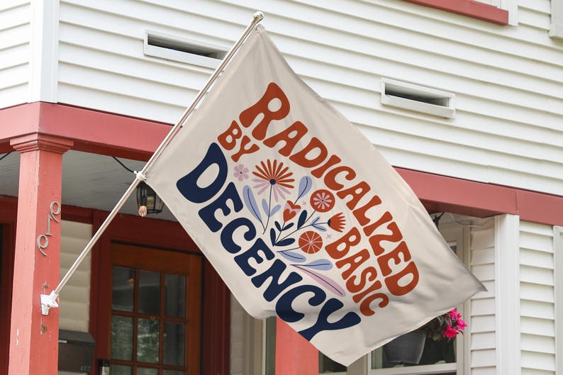 May include: A cream-coloured flag displaying the text "RADICALIZED BY BASIC DECENCY" in orange and navy blue, with floral designs. The flag is mounted outside a house with red trim.
