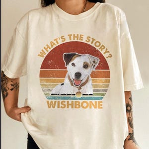May include: A cream-colored t-shirt featuring a graphic of a Jack Russell Terrier dog. The design includes a sunset-colored circle with the text "WHAT'S THE STORY?" above the dog and "WISHBONE" below, all in a distressed, vintage-style font.