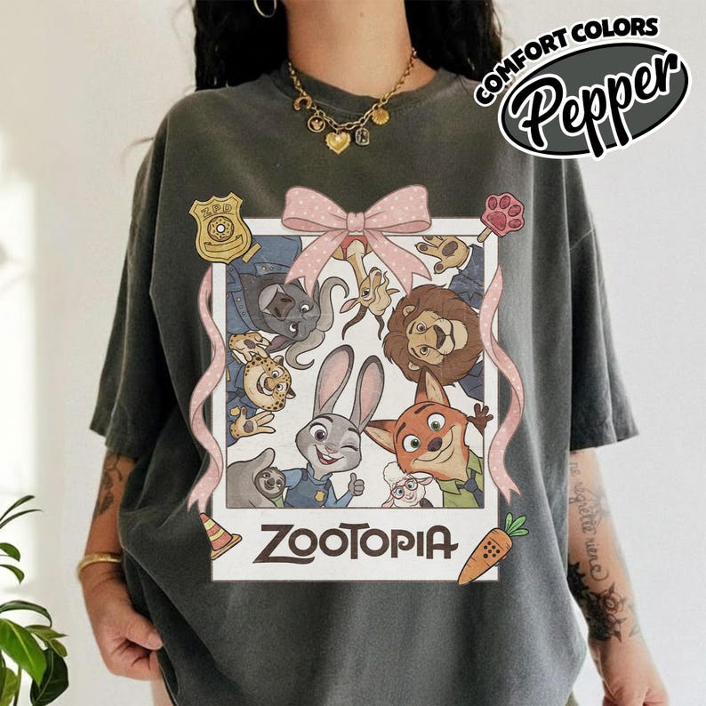 May include: A charcoal gray t-shirt featuring a graphic print of a Polaroid-style frame containing Zootopia characters including Judy Hopps and Nick Wilde. The design includes a pink bow, a ZPD badge, and the text Zootopia at the bottom.