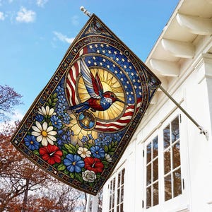 May include: A rectangular flag with a stained-glass design featuring a hummingbird, American flag elements, and floral accents. The flag is mounted on a white building with a blue sky background.