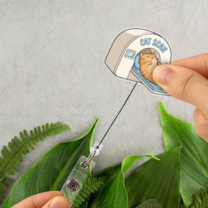 May include: A sticker featuring a cartoon cat inside a CAT SCAN machine. The cat is orange and white, lying on a blue bed. The sticker is held by a hand, with a retractable badge reel attached. Green leaves and ferns are in the background.