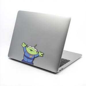 May include: A silver laptop with a cartoon sticker of a green alien character. The alien has three eyes, a blue uniform, and is raising its arms. The laptop is on a white background.