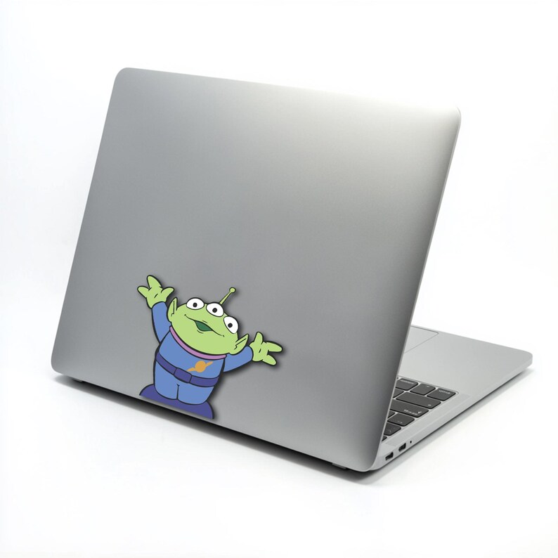 May include: A silver laptop with a cartoon sticker of a green alien character. The alien has three eyes, a blue uniform, and is raising its arms. The laptop is on a white background.