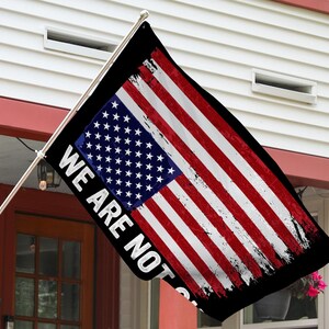 May include: A black flag with a distressed American flag design and the text "We Are Not" printed on it. The flag is hanging from a pole in front of a white house.