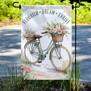 May include: A garden flag featuring a watercolor illustration of a vintage bicycle with flower baskets. The flag has the words "FLOURISH • DREAM • THRIVE" at the top. The bicycle is light green with brown accents, and the flowers are pink and white.