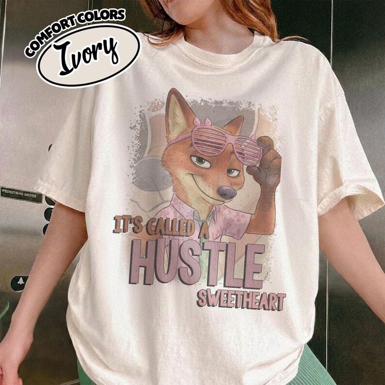 Puede incluir: Una camiseta de manga corta color marfil con un gr&aacute;fico de un zorro con gafas de sol de persiana rosas y una camisa rosa claro. El texto dice IT'S CALLED A HUSTLE SWEETHEART. Una etiqueta en la esquina superior izquierda muestra COMFORT COLORS IVORY.