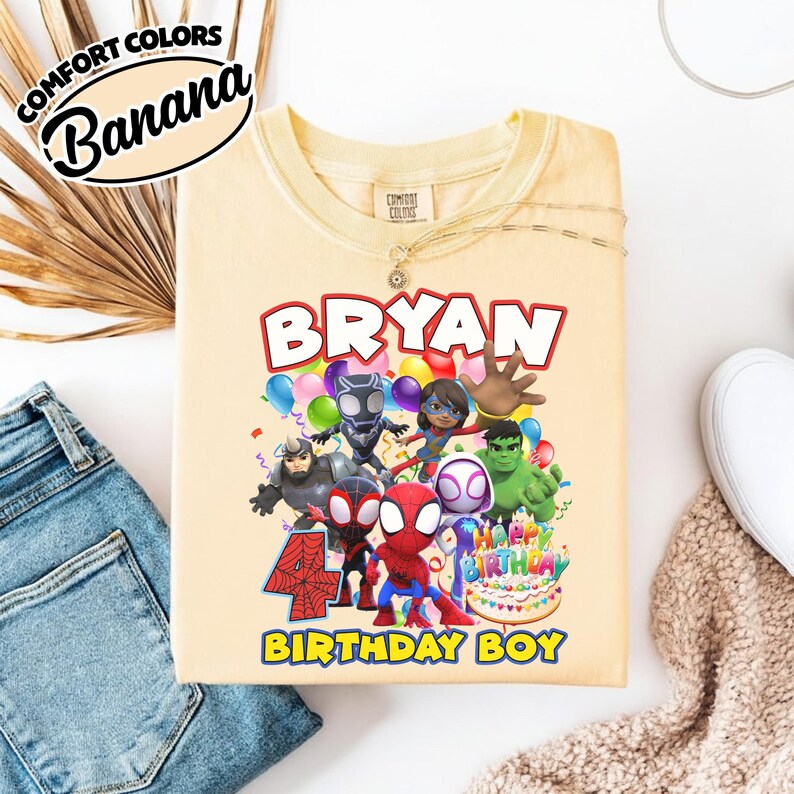 May include: A light yellow t-shirt featuring a graphic print of various superhero characters, the name BRYAN, the number 4 with a spiderweb pattern, and the text HAPPY BIRTHDAY BIRTHDAY BOY, all arranged around a birthday cake illustration.