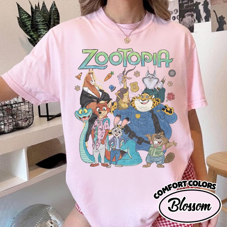 May include: A light pink short-sleeve t-shirt featuring a large graphic print of Zootopia characters including Nick Wilde, Judy Hopps, and Chief Bogo. The text Zootopia is printed in a stylized font above the colorful ensemble of animated animals.