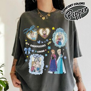 May include: A charcoal gray t-shirt featuring a graphic print of characters from Frozen. The design includes heart-shaped and rectangular frames with illustrations of the characters, blue bows, stars, snowflakes, and the text Birthday girl in white.