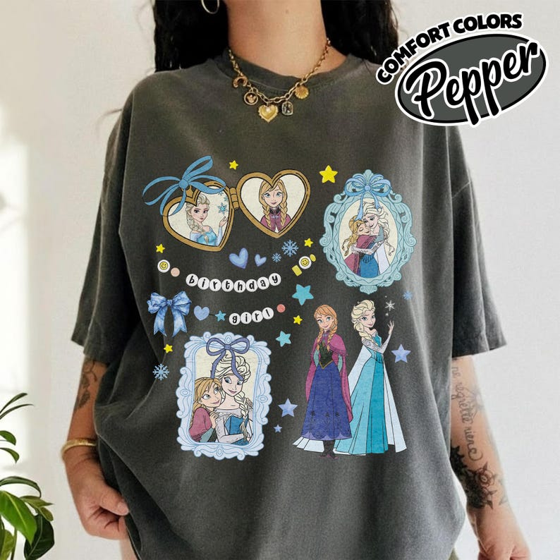 May include: A charcoal gray t-shirt featuring a graphic print of characters from Frozen. The design includes heart-shaped and rectangular frames with illustrations of the characters, blue bows, stars, snowflakes, and the text Birthday girl in white.