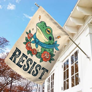 May include: A beige flag with a green frog illustration wearing a blue scarf and surrounded by red roses. The word "RESIST" is printed in dark blue, stylized letters. The flag is mounted on a metal pole against a white house and blue sky.