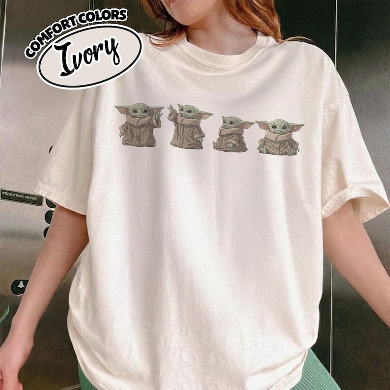 Puede incluir: Una camiseta de manga corta color marfil con un estampado gr&aacute;fico de cuatro peque&ntilde;as criaturas verdes con orejas grandes y t&uacute;nicas color canela en varias poses. El texto COMFORT COLORS IVORY est&aacute; impreso en un &oacute;valo negro arriba a la izquierda.