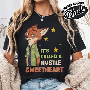 Puede incluir: Una camiseta negra de manga corta con el gr&aacute;fico de un zorro de dibujos animados con camisa verde y corbata morada. El texto dice IT'S CALLED A HUSTLE SWEETHEART, junto a iconos de estrellas amarillas. Etiqueta Comfort Colors.