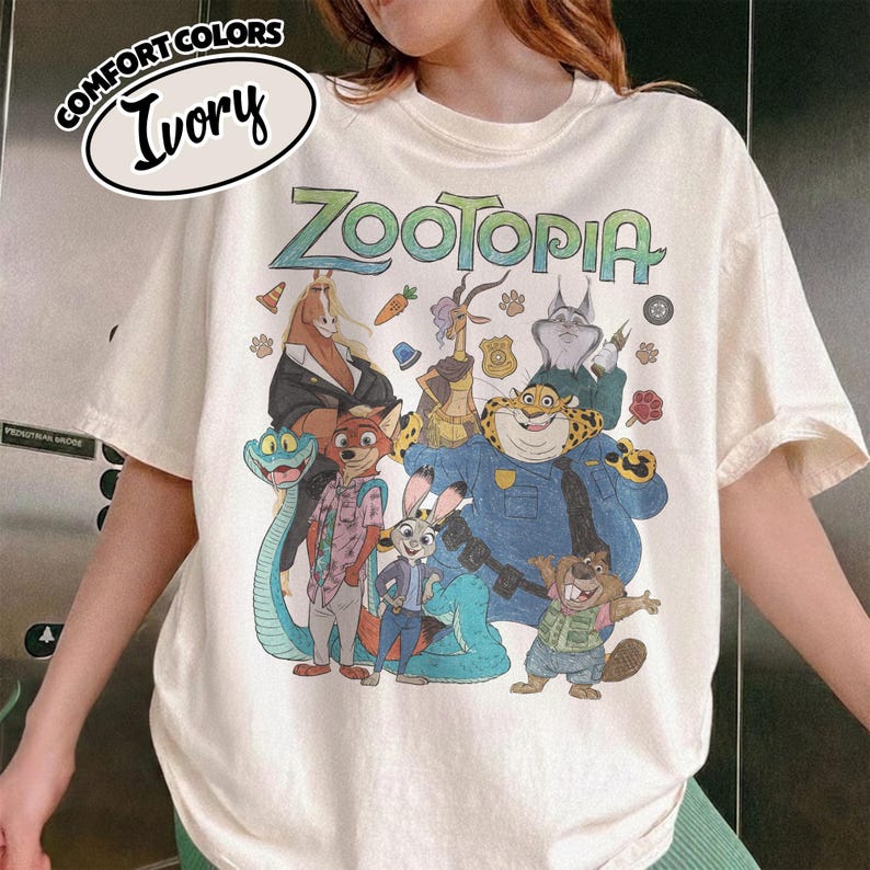 May include: An ivory short-sleeve t-shirt featuring a graphic print of various Zootopia characters including Judy Hopps, Nick Wilde, and Chief Bogo. The text "ZOOTOPIA" is printed in a stylized green font at the top. A label reads "COMFORT COLORS IVORY".