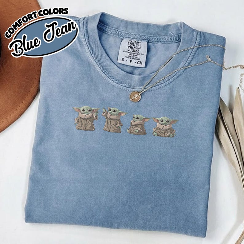 Puede incluir: Una camiseta azul de manga corta con cuello redondo que presenta un gr&aacute;fico bordado de cuatro peque&ntilde;as criaturas verdes con t&uacute;nicas de color canela. La prenda tiene una etiqueta de la marca Comfort Colors y un collar dorado sobre ella.