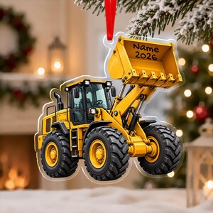 May include: A yellow construction loader ornament hanging from a red ribbon. The vehicle features large black tires, a cab with windows, and a raised front bucket with the text Name 2026 printed in black. The ornament has a clear acrylic border.