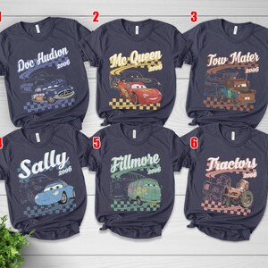 May include: Six dark gray short-sleeve t-shirts featuring graphic prints of characters from the movie Cars. Each shirt displays a character name, the year 2006, and a checkered flag pattern. The characters include Doc Hudson, Mc-Queen, Tow Mater, Sally, Fillmore, and Tractors.