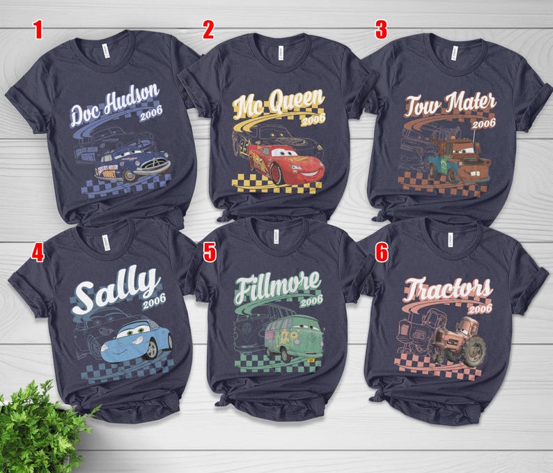May include: Six dark gray short-sleeve t-shirts featuring graphic prints of characters from the movie Cars. Each shirt displays a character name, the year 2006, and a checkered flag pattern. The characters include Doc Hudson, Mc-Queen, Tow Mater, Sally, Fillmore, and Tractors.