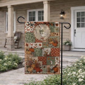 Pu&ograve; includere: Una bandiera da giardino rettangolare con un design patchwork a tema autunnale. Il motivo presenta illustrazioni di oche bianche con fiocchi verdi, zucche arancioni, ghiande marroni e foglie autunnali nei toni del verde, dell'arancione e del marrone.