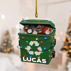 May include: A green Christmas ornament shaped like a recycling bin, filled with cans, bottles, and a gift box. The bin has a white recycling symbol and the name "LUCAS" at the bottom. It's decorated with colorful string lights and a gold hanging cord.