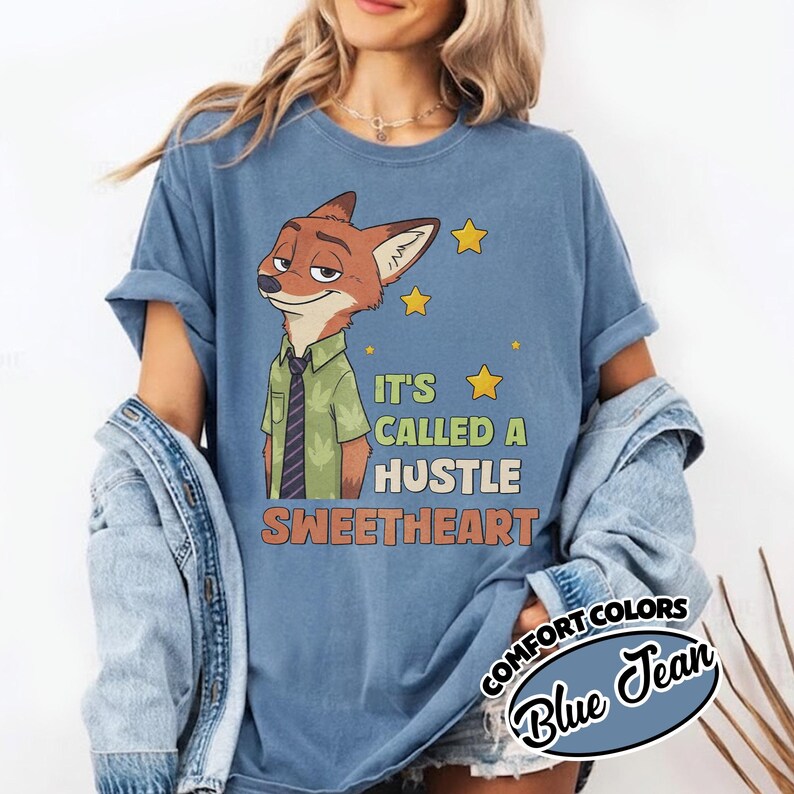 Puede incluir: Una camiseta azul color mezclilla con el gr&aacute;fico de un zorro de dibujos animados con camisa verde y corbata. El texto dice IT'S CALLED A HUSTLE SWEETHEART con estrellas amarillas. Una chaqueta de mezclilla est&aacute; sobrepuesta y una etiqueta circular dice COMFORT COLORS BLUE JEAN.