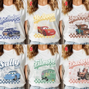 May include: A collection of six white t-shirts featuring Disney Pixar Cars characters. Each shirt displays a character name, the year 2006, a checkered pattern, and a graphic of Doc Hudson, Lightning McQueen, Tow Mater, Sally, Fillmore, or Tractors.