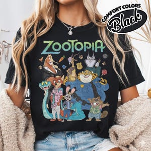 May include: A black short-sleeve t-shirt featuring a graphic print of Zootopia characters including Nick Wilde, Judy Hopps, and Chief Bogo. The text Zootopia is printed in a stylized font above the characters. A Comfort Colors Black label is visible.