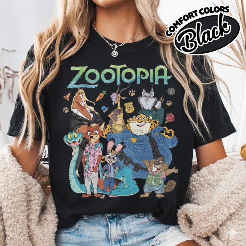 May include: A black short-sleeve t-shirt featuring a graphic print of Zootopia characters including Nick Wilde, Judy Hopps, and Chief Bogo. The text Zootopia is printed in a stylized font above the characters. A Comfort Colors Black label is visible.