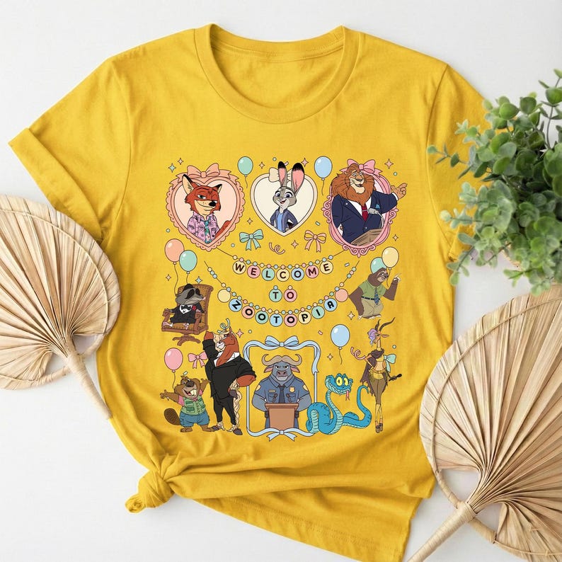 K&ouml;nnte beinhalten: Senfgelbes T-Shirt mit einer farbenfrohen Grafik von Zootopia-Charakteren. Das Design enth&auml;lt den Text "WELCOME TO ZOOTOPIA" und verschiedene Tierfiguren in Rahmen und um den Text herum. Das Shirt ist aus weichem Material.