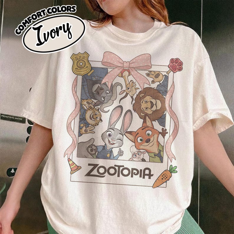 May include: An ivory short-sleeve t-shirt featuring a graphic print of Zootopia characters inside a polaroid-style frame. The design includes a pink bow, a police badge, a traffic cone, and a carrot. The text Zootopia is printed below the frame.