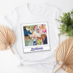 May include: White t-shirt featuring a colorful graphic of characters from the animated film "Zootopia" in a polaroid-style frame. The word "Zootopia" is printed in a stylized font at the bottom of the frame.