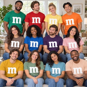 May include: A group of twelve people wearing various colored t-shirts, each featuring a large white lowercase letter m printed on the chest. The shirts are in shades of green, red, yellow, orange, brown, blue, purple, pink, gold, sage, teal, and peach.