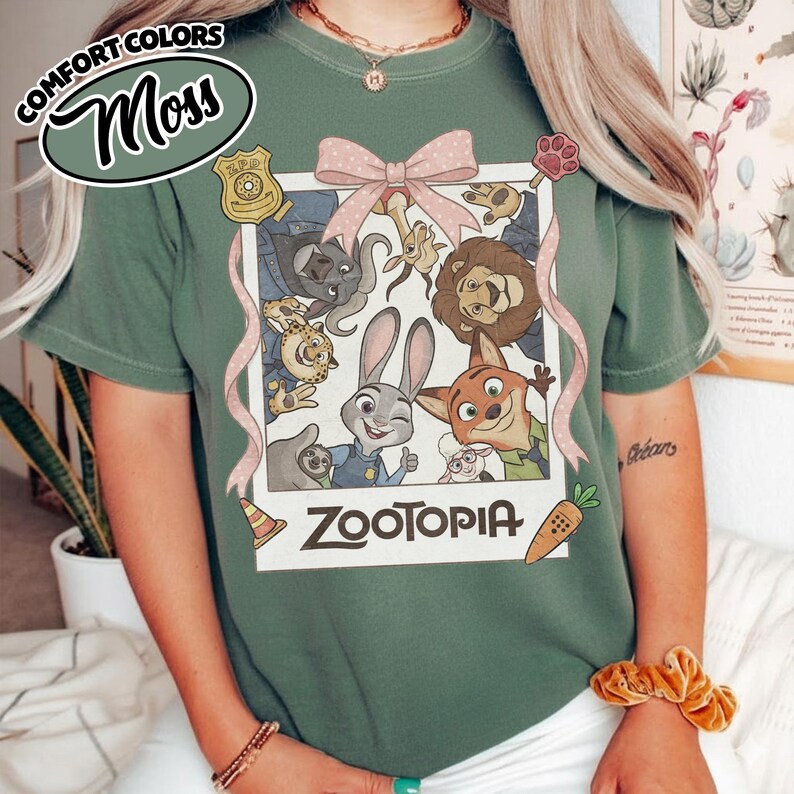 May include: A moss green t-shirt featuring a graphic print of Zootopia characters inside a white polaroid-style frame. The design includes a pink bow, a police badge, a paw print, a traffic cone, and a carrot, with the text Zootopia below.