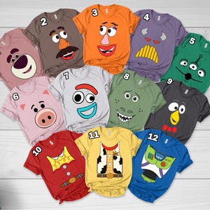 May include: A collection of twelve short-sleeved t-shirts in various colors, each featuring a graphic print of a character face or costume design from the Toy Story franchise, numbered one through twelve and arranged in a heart-shaped layout.