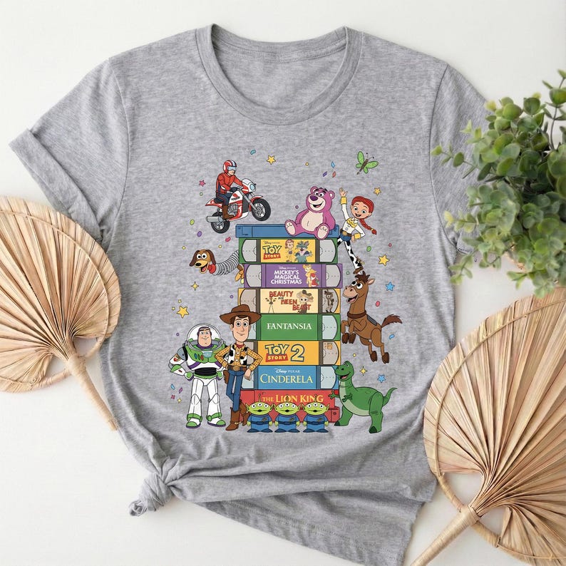 Może przedstawiać: Jasnoszara koszulka z kolorową grafiką postaci z Toy Story i innych animowanych postaci. Projekt zawiera stos kaset VHS z tytułami film&oacute;w i r&oacute;żnymi postaciami.