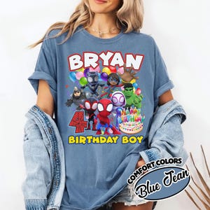 May include: A blue jean colored t-shirt featuring a graphic print of various superhero characters, colorful balloons, a birthday cake, and the text "BRYAN" at the top and "4 BIRTHDAY BOY" at the bottom. The shirt has short sleeves and a crew neckline.