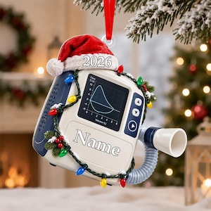 May include: A medical ventilator ornament featuring a red Santa hat, a screen displaying a flow-volume loop graph, and the text 2026 and Name. The device is wrapped in a string of colorful Christmas lights with small pine branches.