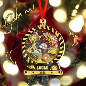 May include: A personalized Christmas ornament with a construction theme. The ornament is round with a black and yellow border, featuring construction vehicles, hard hats, and the name "LUCAS" and the year "2025". It has a red ribbon for hanging and the text "Shake me!"