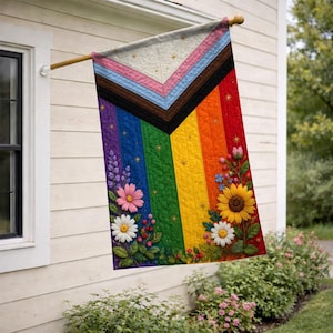 May include: A vertical quilted flag featuring the Progress Pride flag design with a chevron pattern in white, pink, light blue, brown, and black, overlaid on a rainbow background. The flag is decorated with embroidered sunflowers and daisies.