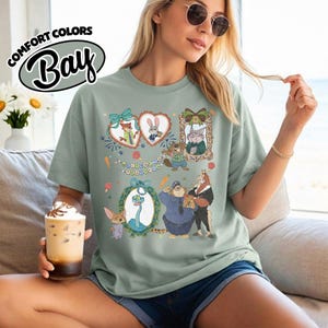 May include: A sage green short-sleeve t-shirt featuring a graphic print of various animated animal characters from Zootopia, including a fox, rabbit, and sloth, framed in decorative hearts and ovals with the text Welcome to Zootopia.