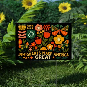 May include: A black yard sign with a colorful floral design and butterflies. The sign reads "IMMIGRANTS MAKE AMERICA GREAT" in yellow and white text. The sign is displayed on a metal stake in a grassy yard.