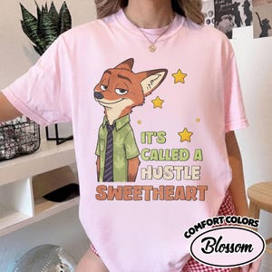 Puede incluir: Una camiseta rosa claro con el gr&aacute;fico de un zorro de dibujos animados con camisa verde y corbata oscura. El texto dice IT'S CALLED A HUSTLE SWEETHEART en letras verdes y naranjas, acompa&ntilde;ado de estrellas amarillas. Tiene cuello redondo.