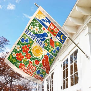 May include: A rectangular flag with a colorful, illustrated design. The flag features the text "HATE HAS NO HOME HERE" and various images, including birds, flowers, a house, and a sun. The flag is hanging from a metal pole attached to a white house.